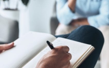 Close-up shot of a person writing with a pen and a pad with another person sitting in the background.